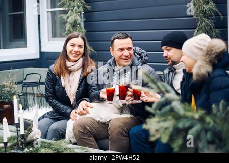 Lagerfeuer-Party. Fröhliches junges Familienpaar, das sich im Winter mit Freunden am Lagerfeuer versammelt, glückliche Menschen, die heißen Glühwein zubereiten und trinken Stockfoto