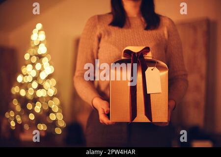 Weihnachtsgeschenk. Unbekannte Frau hält große goldene Weihnachtsschachtel mit roter Schleife und steht vor verschwommenem Hintergrund des Weihnachtsbaums, selektiv Stockfoto