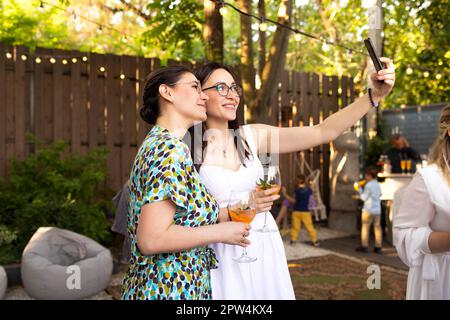 Glückliche Freundinnen verbringen Zeit miteinander, junge Frau trinkt Aperol Spritz Cocktail auf einer Hochzeitsfeier im Freien, macht Selfi zusammen. Glück Stockfoto