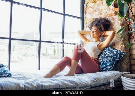 Ein entspanntes, afroamerikanisches Mädchen in Hausbekleidung, das auf einer Fensterbank sitzt, eine glückliche Afro-Frau zu Hause, die sanft zum Fenster lächelt Stockfoto