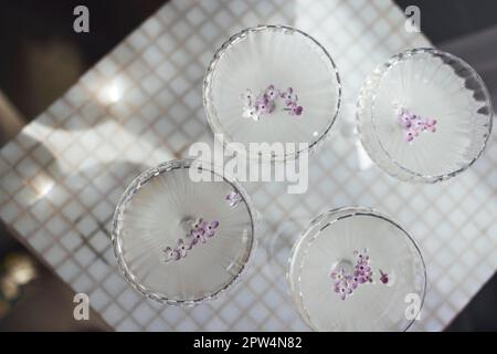 Vier Gläser mit Champagner auf einer hellen Tischoberfläche, die den weichen Sonnenschein reflektiert, rayson. Sekt mit Fliederblüten. Draufsicht Stockfoto