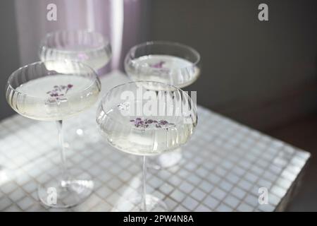 Vier Gläser mit Champagner auf einer hellen Tischoberfläche, die den weichen Sonnenschein reflektiert, rayson. Sekt mit Fliederblüten. Draufsicht Stockfoto