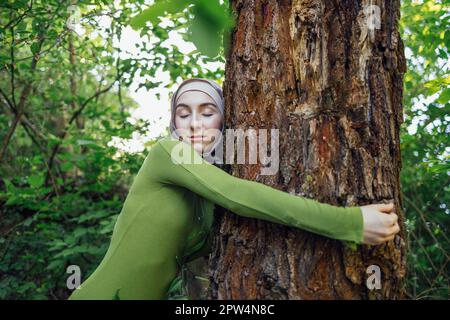 Muslimisches Teenager-Mädchen, das einen Baum umarmt. Nahaufnahme eines jungen Weibchens in einem Hijab und Freizeitkleidung im Park. Umweltschutz und Fürsorge Stockfoto