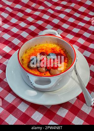 Creme Brulee, verbrannte Sahne. Süßes gebackenes Dessert auf einem weißen Porzellanteller mit Griffen. Gericht, dekoriert mit Johannisbeeren und Blaubeeren. Platte und Stockfoto