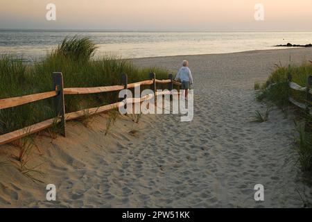 Eine Seniorin geht allein auf einem Pfad durch die Dünen zum Meer bei Sonnenuntergang Stockfoto