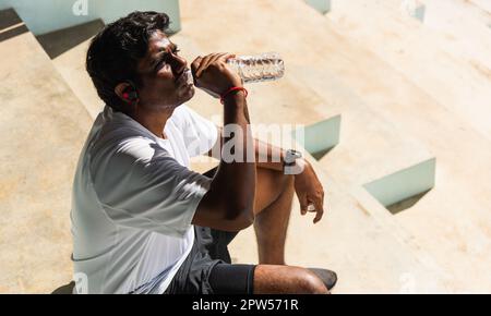 Nahaufnahme Asian junge Sportlerin schwarzer Mann tragen Athlet Kopfhörer er trinkt Wasser aus einer Flasche nach dem Laufen im Outdoor-Street Health Park, h Stockfoto