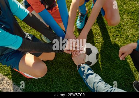 Der Fußballtrainer motiviert die kleine Fußballmannschaft, bevor er die Hände am Ball zusammenstapelt Stockfoto