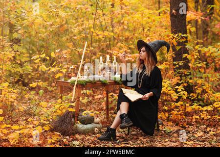 Junge Frau im Hexenkostüm sitzt mit Magie am Tisch Zutaten und Notizen machen am Herbsttag im Wald Stockfoto
