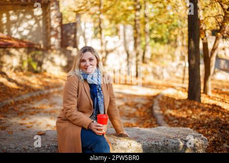 Eine wunderschöne, lächelnde blonde Frau mittleren Alters mit beigefarbenem Mantel und Schal, die am sonnigen Herbsttag im Stadtpark sitzt und ihre Tasse Kaffee zum Mitnehmen genießt. Stockfoto