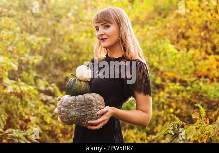 Porträt einer schönen, jungen blonden Frau mit langen, glatten Haaren und dunklem Vampir-Make-up, die in die Kamera schaut, ein schwarzes Guipure-Kleid trägt, hält Stockfoto