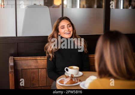 Zwei entzückende Freundinnen, die Kaffee trinken und sich in einem gemütlichen Café unterhalten, lächelnde weibliche beste Freundinnen, die eine angenehme Unterhaltung beim Treffen in genießen Stockfoto