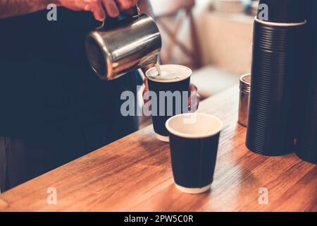 Crop Barista Gießen heiße Milch aus dem Eisbecher in Kaffeetassen. Ein großer anonymer Profi, der Cappuccino und Latte zum Mitnehmen zubereitet Stockfoto