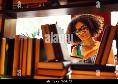Glückliche afroamerikanische Studentin, die in der Universitätsbibliothek studding und auf der Suche nach dem Buch ist, mit Brille Stockfoto