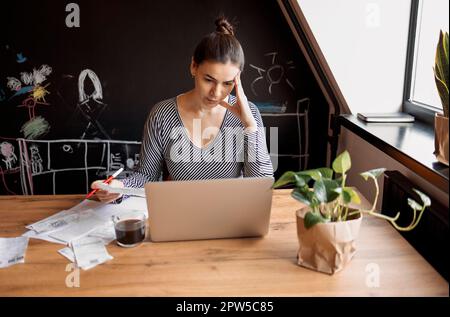 Seriöse Hausfrau sitzt am Tisch in der Küche, prüft die Haushaltsrechnungen, macht Berechnungen mit dem Taschenrechner, bezahlt Dienstprogramme über das E-Banking-System ein Stockfoto