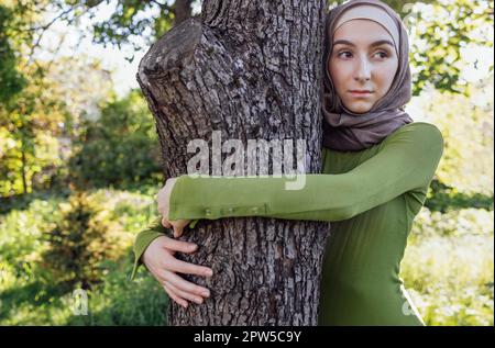 Muslimisches Teenager-Mädchen, das einen Baum umarmt. Nahaufnahme eines jungen Weibchens in einem Hijab und Freizeitkleidung im Park. Umweltschutz und Fürsorge Stockfoto