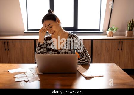 Seriöse Hausfrau sitzt am Tisch in der Küche, prüft die Haushaltsrechnungen, macht Berechnungen mit dem Taschenrechner, bezahlt Dienstprogramme über das E-Banking-System ein Stockfoto