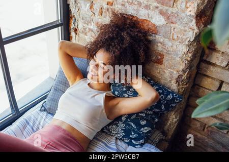 Ein entspanntes, afroamerikanisches Mädchen in Hausbekleidung, das auf einer Fensterbank sitzt, eine glückliche Afro-Frau zu Hause, die sanft zum Fenster lächelt Stockfoto