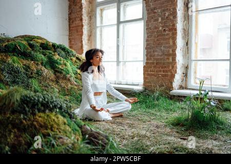 Großes Foto einer optimistischen brünetten Dame, die auf dem Rasen sitzt. Rest tragen Sie weiße bequeme Kleidung, Oberteil und Hose, innen. Ungewöhnlicher Ort. Gesundheitswesen Stockfoto