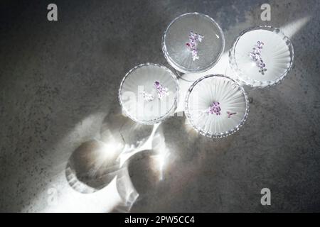 Vier Gläser mit Champagner auf einer hellen Tischoberfläche, die den weichen Sonnenschein reflektiert, rayson. Sekt mit Fliederblüten. Draufsicht Stockfoto