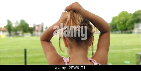 Junge hübsche Frau hat sich die Haare gebunden. Ein Mädchen wird von hinten vor dem Hintergrund eines Fußballfelds fotografiert. Die Bildung des Haarschwanzes. Stockfoto