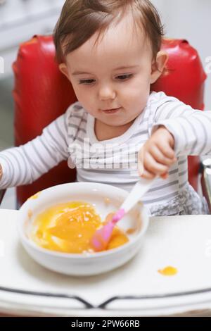 Sie wird immer besser koordiniert. Ein süßes kleines Baby, das in einem Hochstuhl sitzt und isst Stockfoto