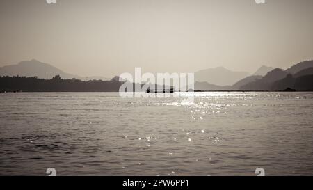 Sepiabarbene Silhouette des Slow Bootes auf dem Mekong, der an Laos und Thailand grenzt, mit Umrissen der umliegenden Berge. Stockfoto