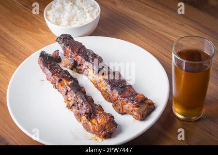 Geröstete Schweinerippchen mit Barbecue-Sauce, Reis und Guarana-Limonade auf einem weißen Porzellanteller. Holztisch. Stockfoto