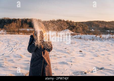Eine Frau mit geschlossenem Gesicht wirft Schnee mit den Händen auf ein schneebedecktes Feld Stockfoto