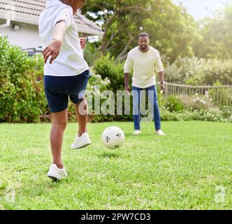 Seine Koordination mit dem Training wird besser. Ein kleiner Junge tritt seinem Vater einen Fußball zu, während er draußen zusammen spielt Stockfoto