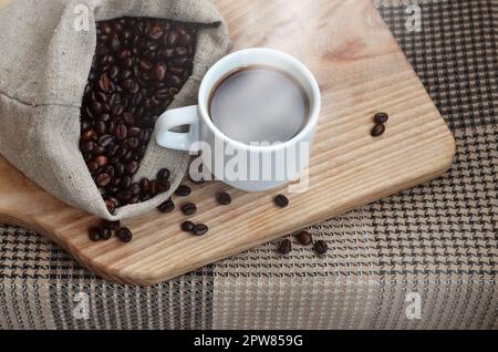 Einen vollen Beutel der braunen Bohnen und eine weiße Tasse heißen Kaffee liegt auf einem Holz- Oberfläche. Attribute, die mit der Zubereitung von Kaffee im Zusammenhang Stockfoto