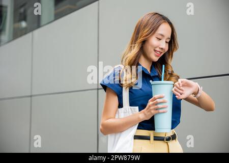 Asiatische schöne Frau zuversichtlich lächelnd mit Stofftasche hält Stahl Thermos Becher Wasser Glas sie zu Fuß im Freien auf der Straße in der Nähe von modernen Gebäude Stockfoto