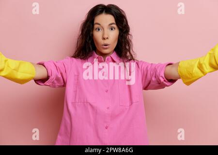 Junge Hausfrau in pinkfarbenem Hemd und gelben Reinigungshandschuhen, hält eine Kamera im Selfie-Modus mit überraschtem Gesicht. Hochwertiges Foto Stockfoto