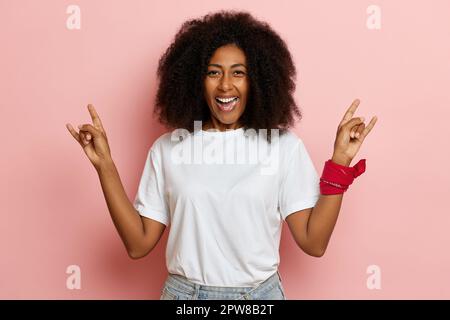 Junge positive lockige, dunkelhaarige Frau lächelt und zeigt Horngeste, sie steht isoliert über rosa Mauern. Hochwertiges Foto Stockfoto