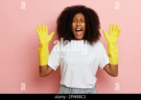 Eine enttäuschte, lockige junge Frau mit weit verbreitetem Mund hält die Handflächen hoch und verärgert sich über die Hausarbeit, trägt Gummischutzhandschuhe zum Reinigen Stockfoto