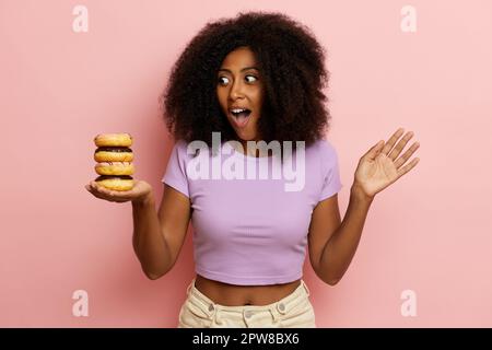 Porträtaufnahme einer überraschten, dunkelhäutigen jungen Frau mit einem überfreulichen Lächeln, leckeren Donuts und einem aufregenden, isolierten Blick auf pinkfarbenen Hintergrund. Stockfoto