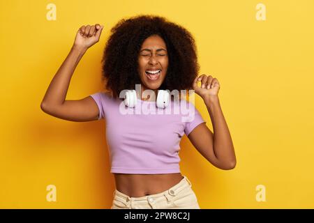 Horizontale Aufnahme einer fröhlichen, lockigen Frau mit erhobenen Händen in der Luft, tanzt gern in Bewegung, trägt weiße, kabellose Kopfhörer, angezogen i Stockfoto
