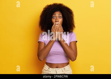 Eine verängstigte, lockige Frau steht an einer gelben Wand, sie hielt sich die Handflächen an den Mund und öffnete ihre Augen weit, als sie einen Schock von riesigem Rabatt hatte. Stockfoto