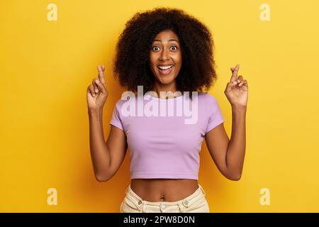 Innenaufnahme einer aufgeregten jungen Frau mit buschiger Frisur und gedrückten Daumen, wünscht sich was, lächelt zahnvoll, trägt ein lila T-Shirt, isoliert über Yello Stockfoto