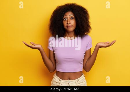 Eine zweifelhafte, lockige, brünette Frau hält die Hände in zögerlichen Gesten, kämpfte um eine Entscheidung, trug lässige Kleidung, isoliert über gelbem Rücken Stockfoto