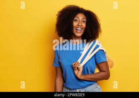 Eine dunkelhäutige, lockige Frau steht neben einer gelben Wand, hält eine Tasche mit Orangenfrüchten und lächelt. Stockfoto