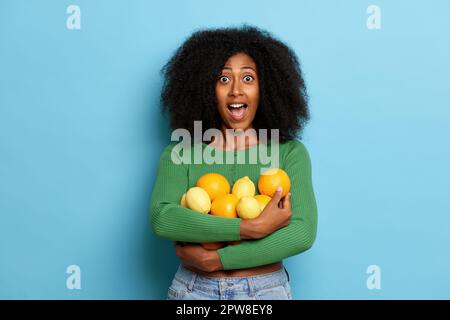 Erstaunliche dunkelhäutige, lockige Frau hält einen Haufen frischer Orangen und Zitronen, sieht aufgeregt aus, trägt grüne Strickjacke und blaue Jeans, isoliert über blauem Rücken Stockfoto
