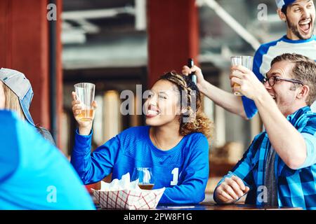 Sie erheben ihre Brille auf ein episches Spiel. Eine Gruppe von Freunden jubelt, während sie ein Sportspiel in einer Bar anschauen. Stockfoto
