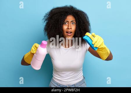 Eine afrikanische Frau mit lockigem Haar, die sich zu Hause auf die Reinigung vorbereitet, hält die Bürste und die Waschflüssigkeit mit einem überraschenden Lächeln in der Hand. Hochwertiges Foto Stockfoto