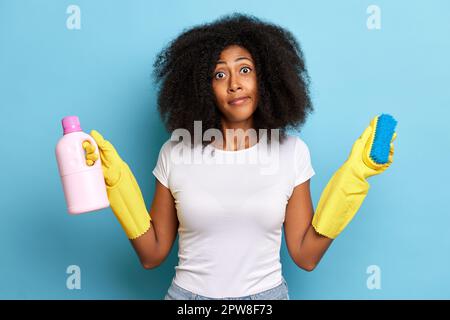 Hausfrau in gelben Gummihandschuhen steht mit Reinigungsausrüstung isoliert auf blauem Hintergrund, sie hält die Bürste und die Waschflüssigkeit. Hohe Qualität Stockfoto