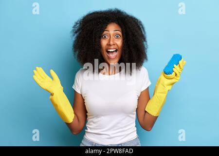 Dunkelhäutige Hausfrau mit überfreulichem Gesichtsausdruck, öffnet den Mund weit, trägt geschützte Handschuhe, hält Reinigungsbürste, isoliert auf blauem Hintergrund. Hoch Stockfoto