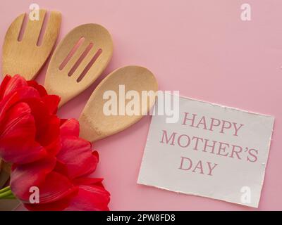 Rote Tulpen blühen mit Küchenutensilien auf rosa Hintergrund mit weißem Papier mit Happy Mütters Day. Grußkarte zum Frauen- und Muttertag. Geburtstag congra Stockfoto