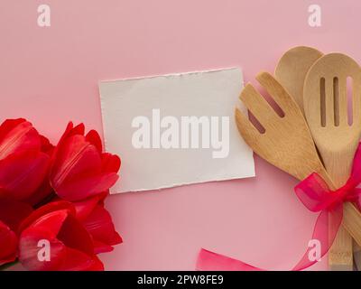 Rote Tulpen mit Küchenutensilien auf pinkfarbenem Hintergrund mit weißem Papier und Kopierbereich. Grußkarte zum Frauen- und Muttertag. Glückwunsch zum Geburtstag Stockfoto