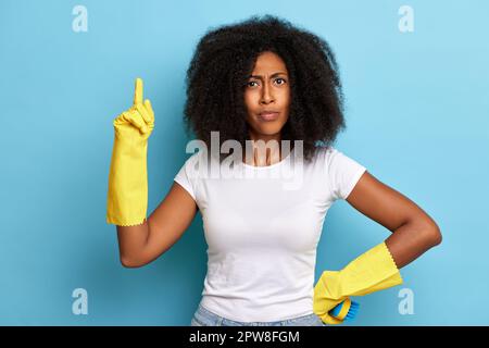 Eine afroamerikanische lockige Frau, die mit dem Finger nach oben zeigt. Sie verkleidete sich als Hausfrau mit gelben Handschuhen. Stockfoto