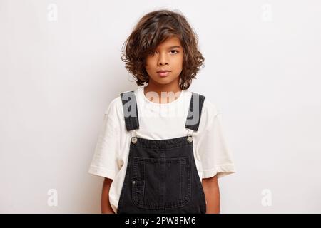 Junger, dunkelhäutiger, charmanter Teenager, der ein weißes T-Shirt und einen schwarzen Overall trägt, steht normalerweise vor weißem Hintergrund. Stockfoto