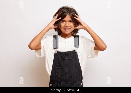 Verärgerter junger Kerl in Jeans Overall, hat Handflächen auf dem Kopf, leidet an Kopfschmerzen, isoliert über weiße Wand. Hochwertiges Foto Stockfoto